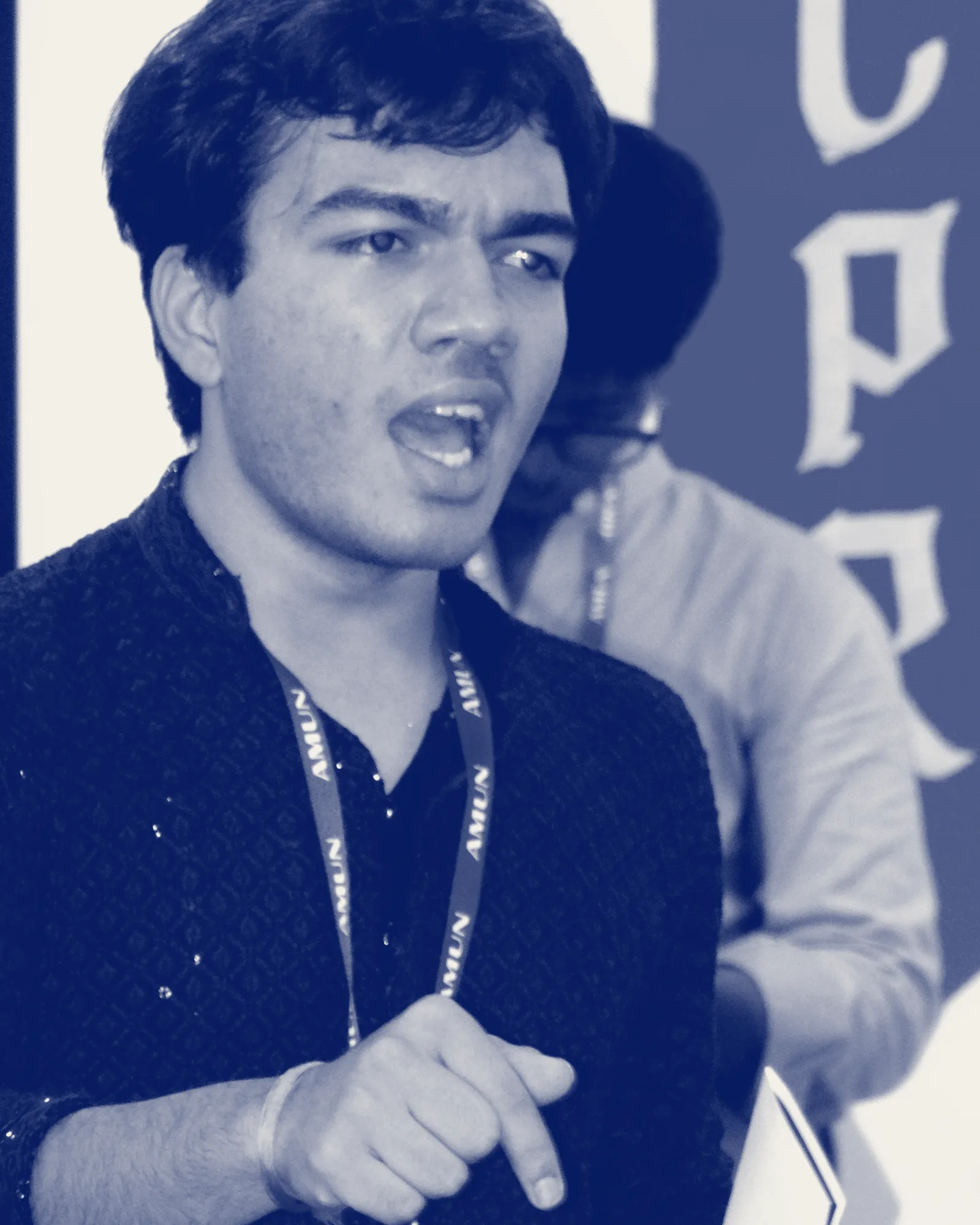 A delegate speaking mid-sentence, pointing, an AMUN lanyard around his neck.